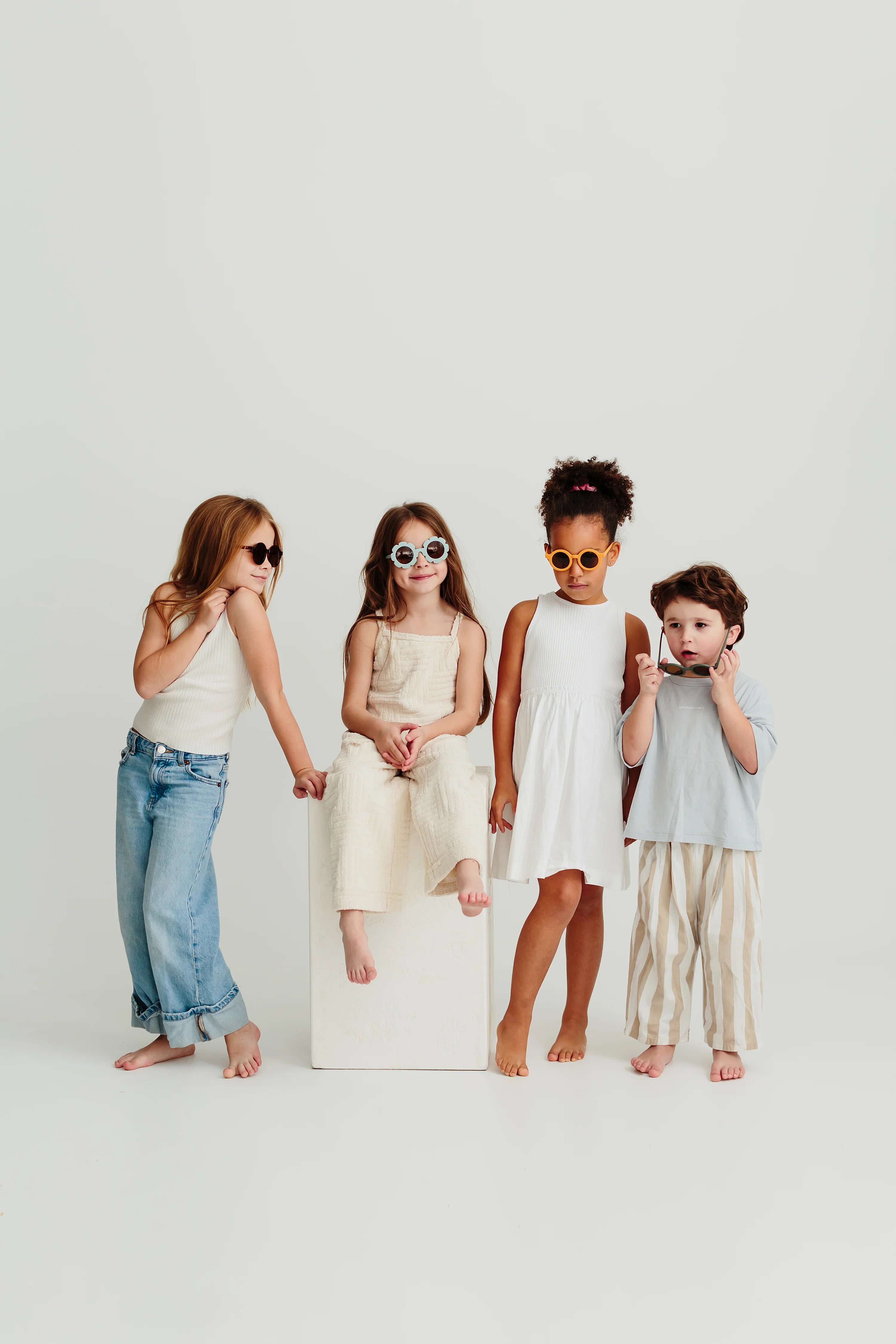 Four children stand together against a plain background, all wearing stylish summer clothes and Kids Shades Sand from LITTLE DROP with UV400 Protection. The child on the far left is striking a pose, while the two center children are sitting and standing on a white block. The child on the right is smiling brightly.