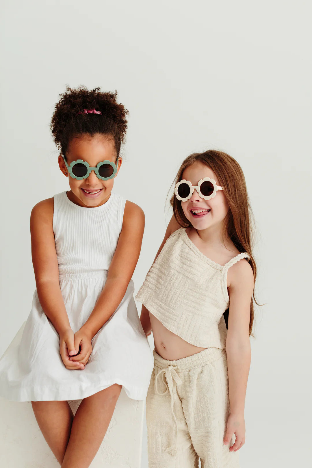 Two young girls smiling at the camera while wearing stylish Daisy Shades Bone by LITTLE DROP, which offer UV400 protection. The girl on the left has curly hair tied back and is dressed in a white sleeveless dress. The girl on the right has long straight hair and is wearing a beige sleeveless top and shorts set, both holding their silicone sunglasses cases.