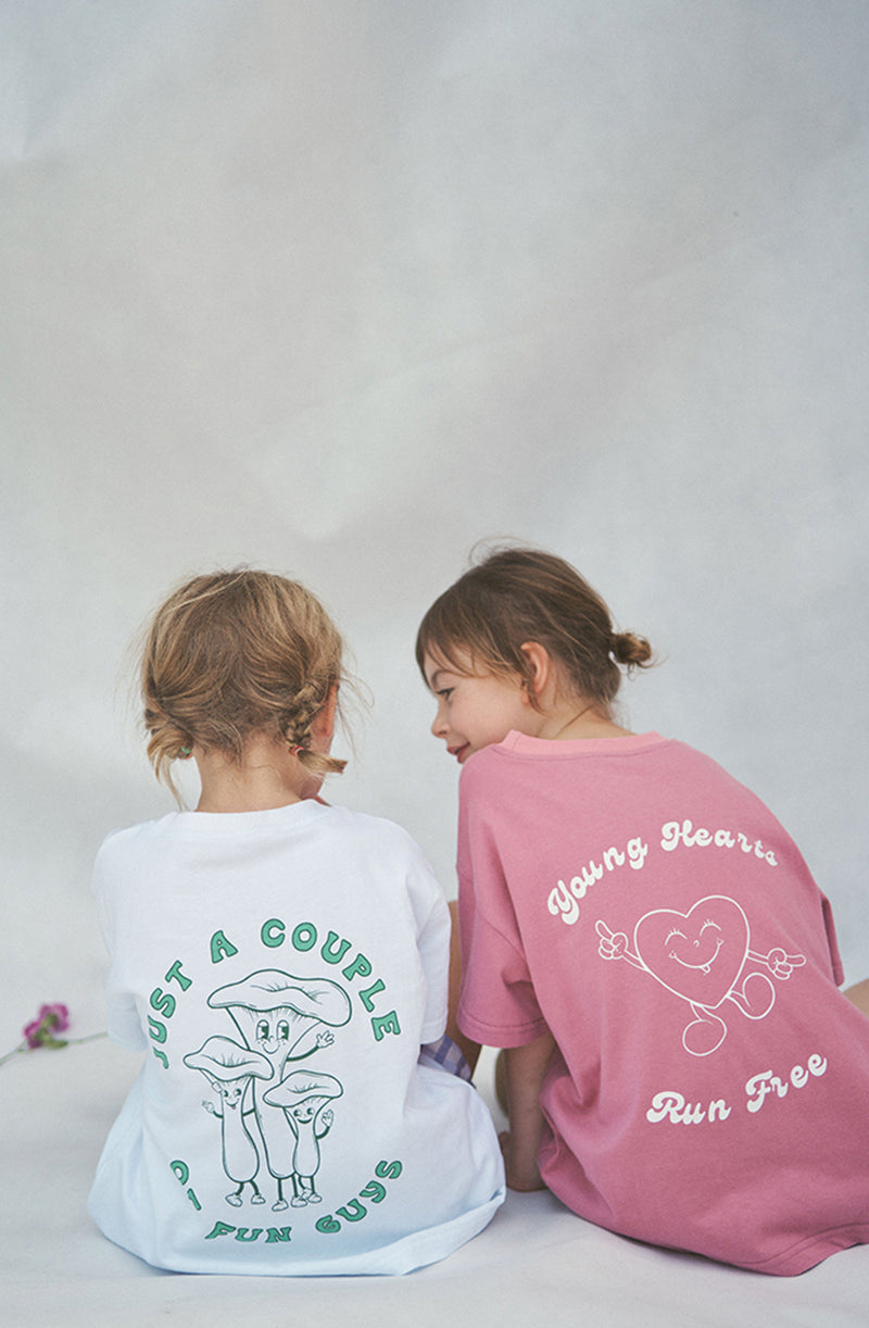 Two girls sitting side by side showing the back prints of Fun Guys tee and Young Hearts tee