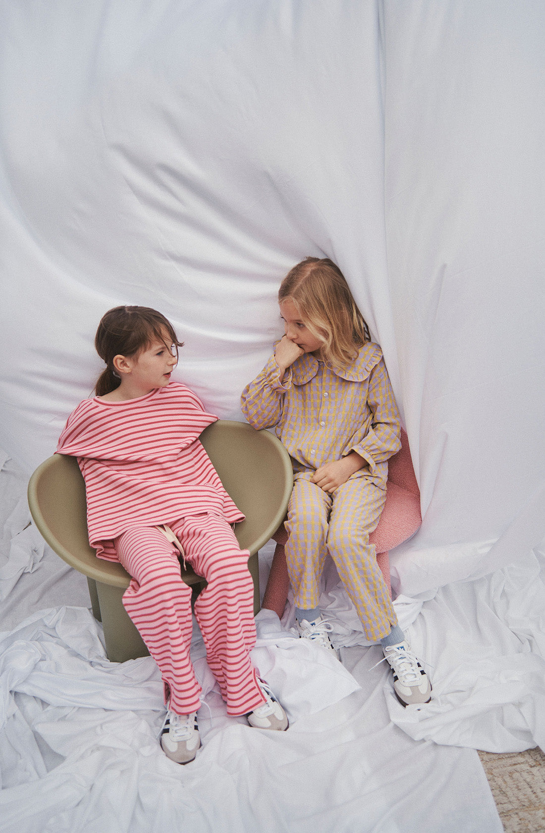 Two girls sitting on chairs in matching sets of gingham and striped outfits.