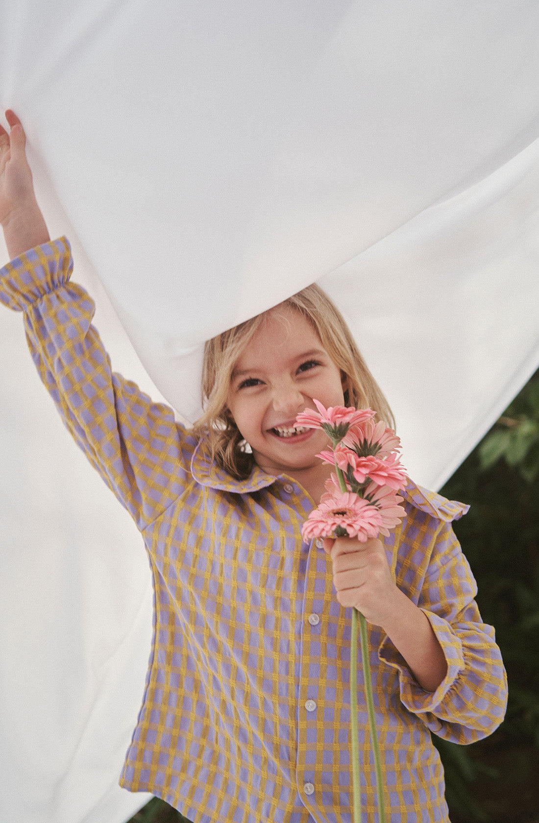 Young kid wearing the Maggie blouse l and matching pant holding flowers under a white sheet.