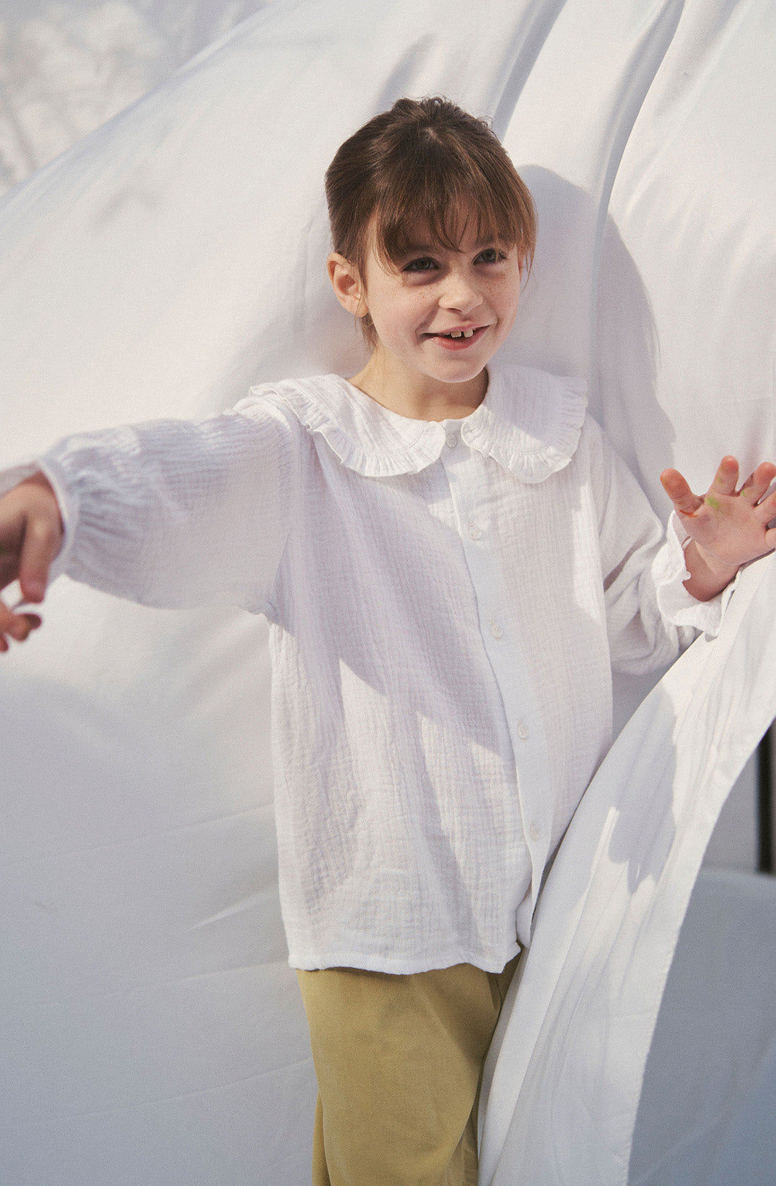 Young kid wearing the Maggie muslin blouse and mustard pants playing under a white sheet.