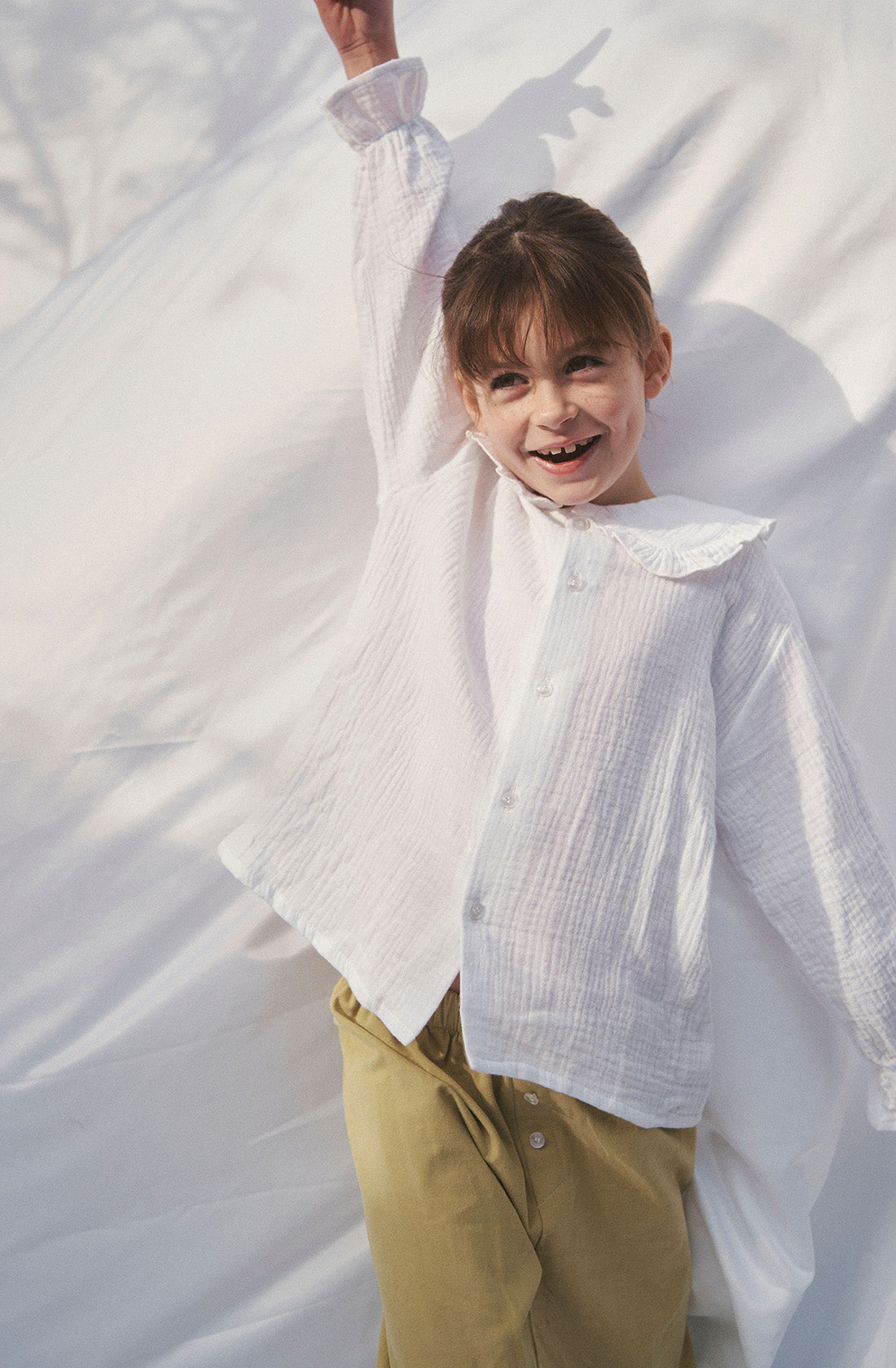 Young kid wearing the Maggie muslin blouse and mustard pants playing under a white sheet.