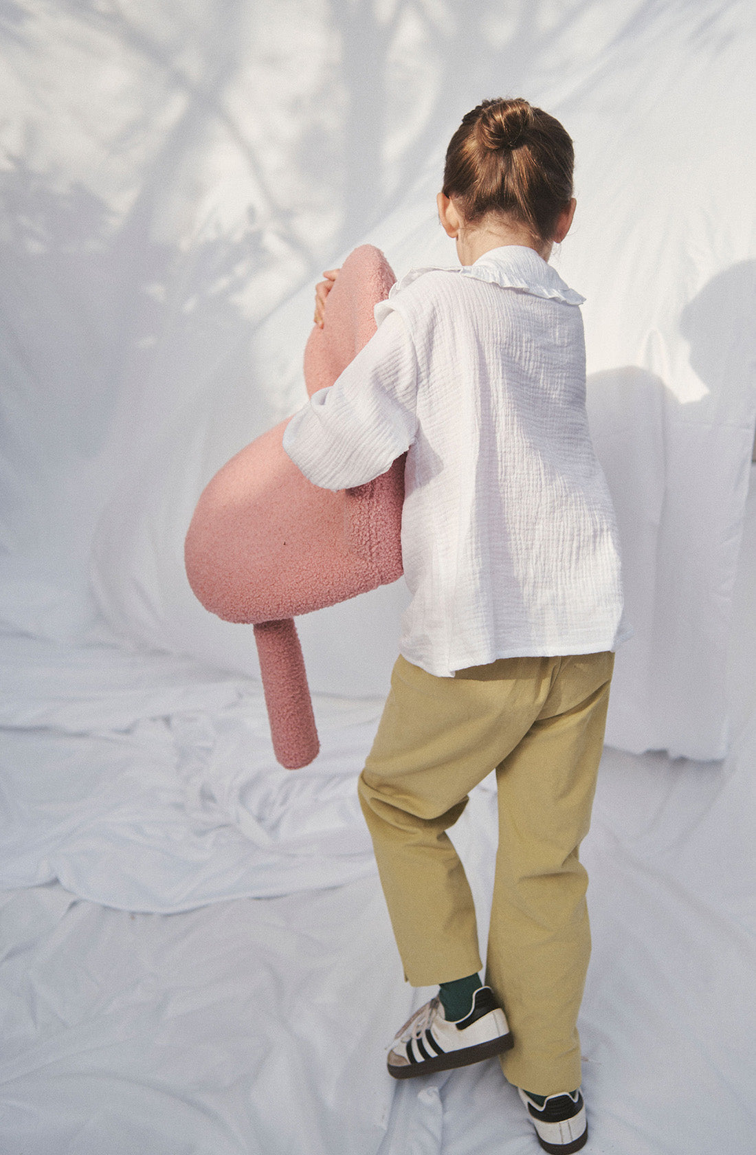 Young kid wearing the Maggie muslin blouse and mustard pants holding a woolie chair.