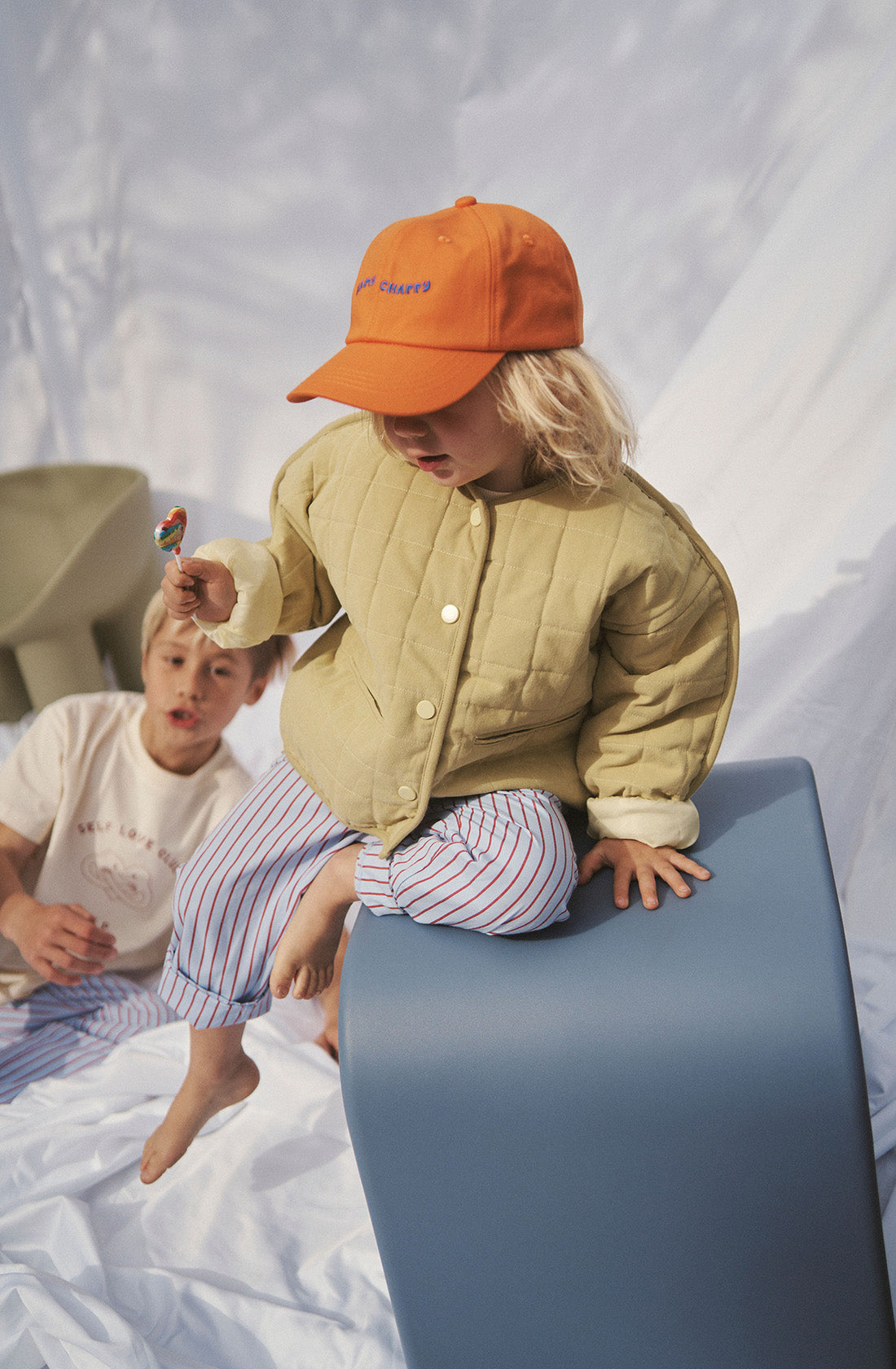 Young kid wearing the parker quilted jacket and striped pants and holding a lollypop while sitting a a blue chair.