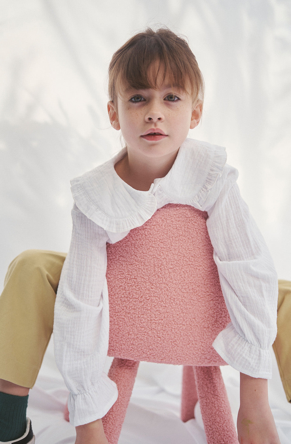 Young kid wearing the Maggie muslin blouse and mustard pants sitting on a chair.