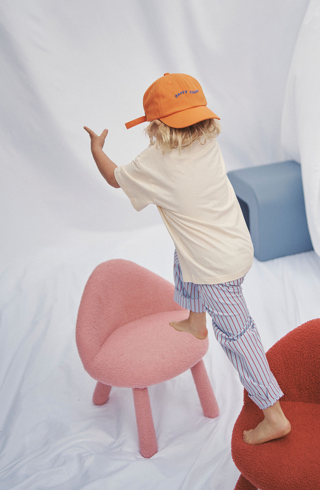 Young kid wearing the party pant blue and red striped in a beige top and orange hat jumping between chairs.