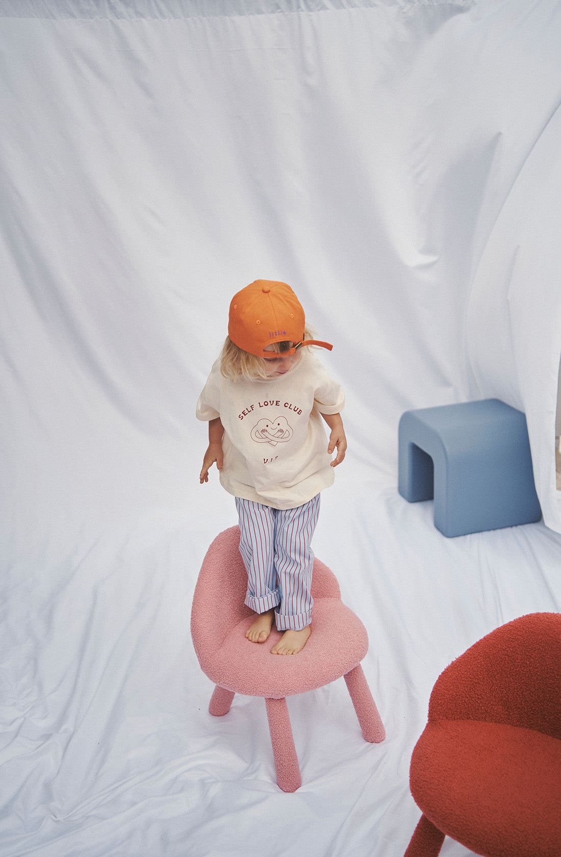 Young kid wearing the party pant blue and red striped in a beige top and orange hat  standing on a chair.