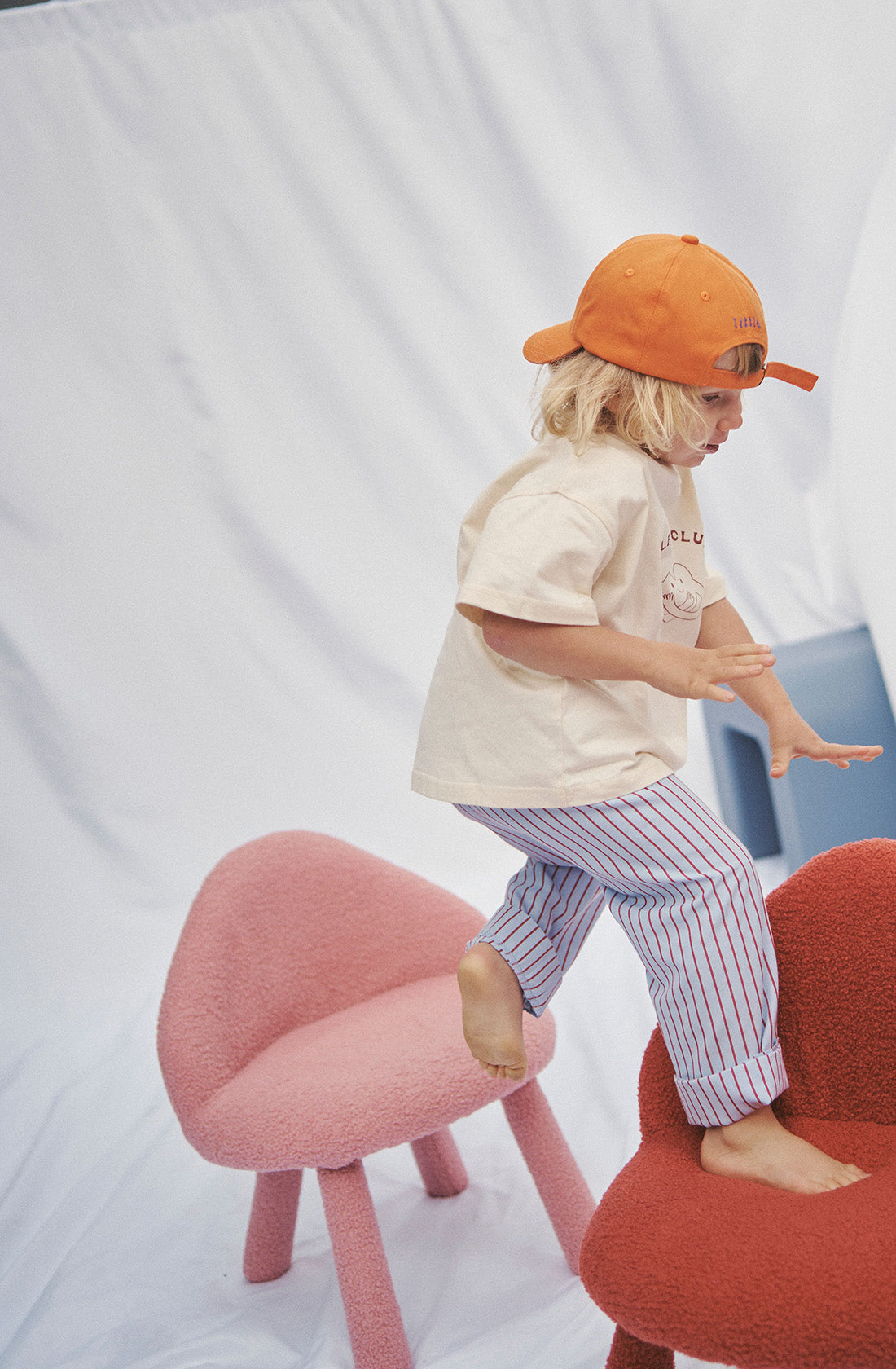 Young kid wearing the party pant blue and red striped with a beige top and orange hat standing on a chair.