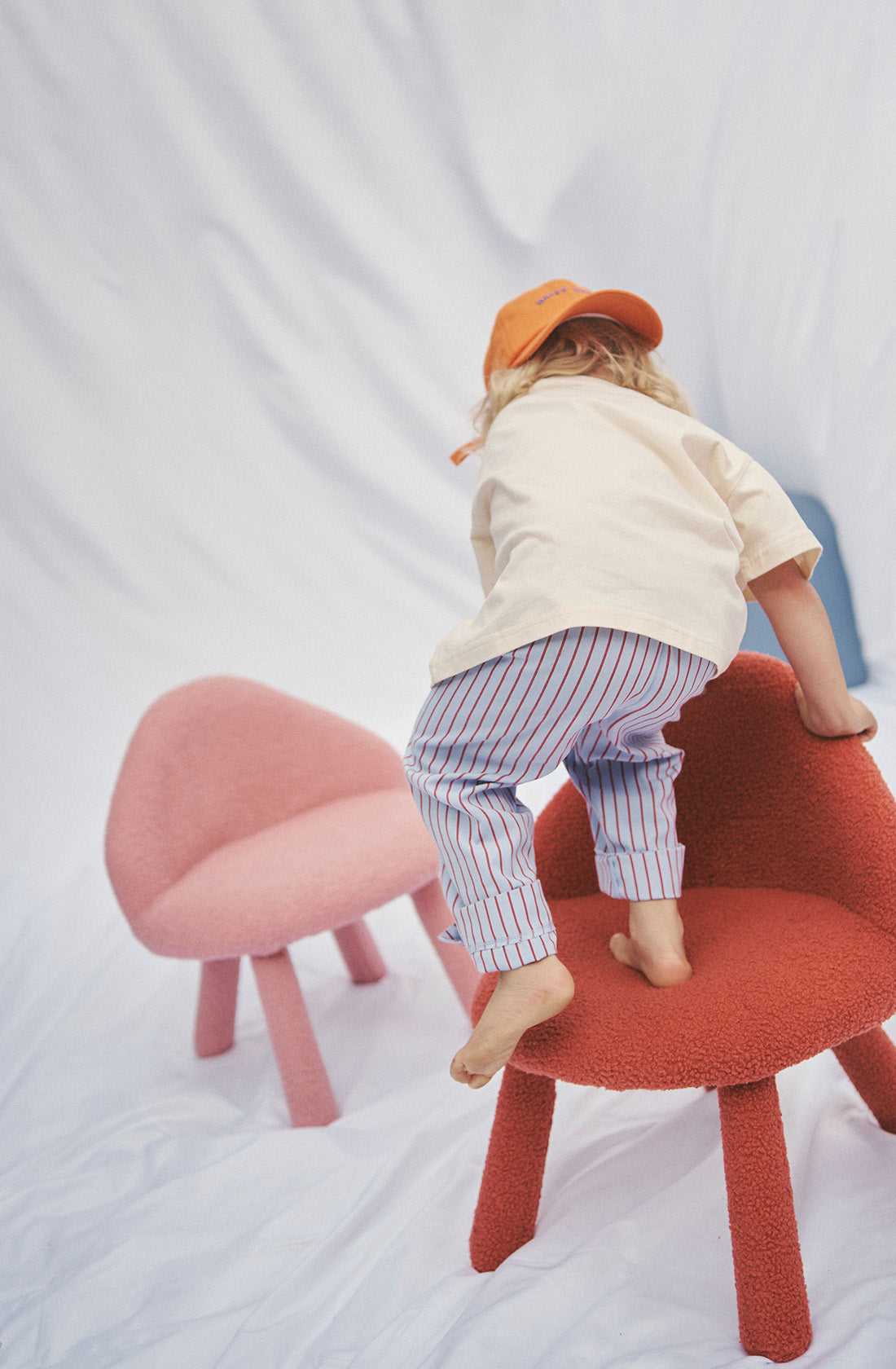 Young kid wearing the party pant blue and red striped in a beige top and orange hat standing on a chair.