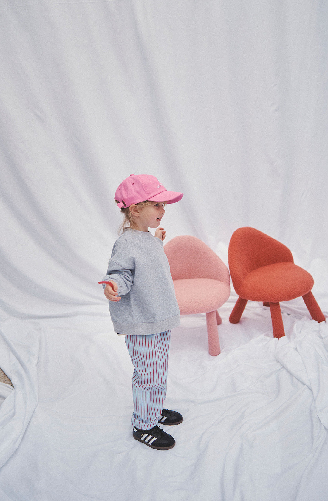 A young girl wearing a grey jumper and striped pants and pink hat. There are two chairs behind her.