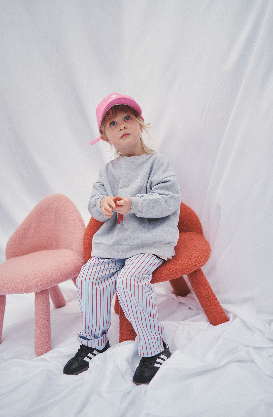 A young girl wearing a grey jumper and striped pants with a pink had sitting on a red chair.