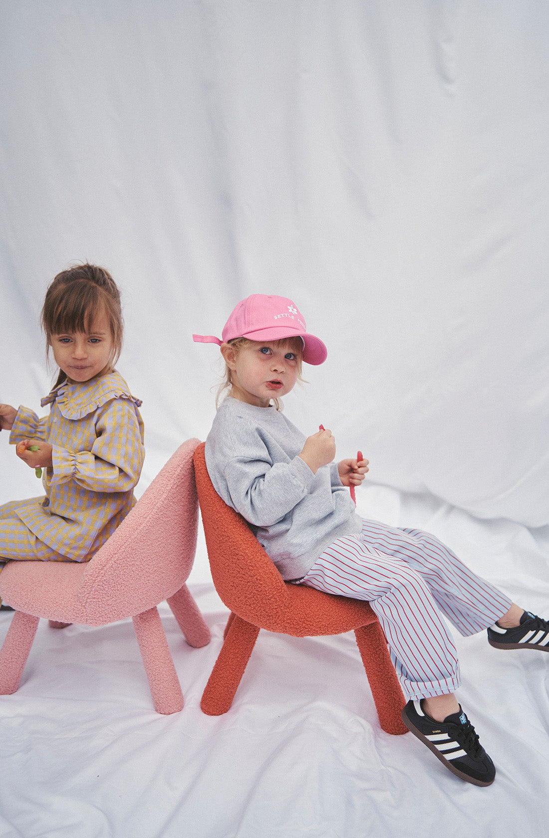 A young girl wearing a grey jumper and striped pants with a pink hat sitting on a chair.