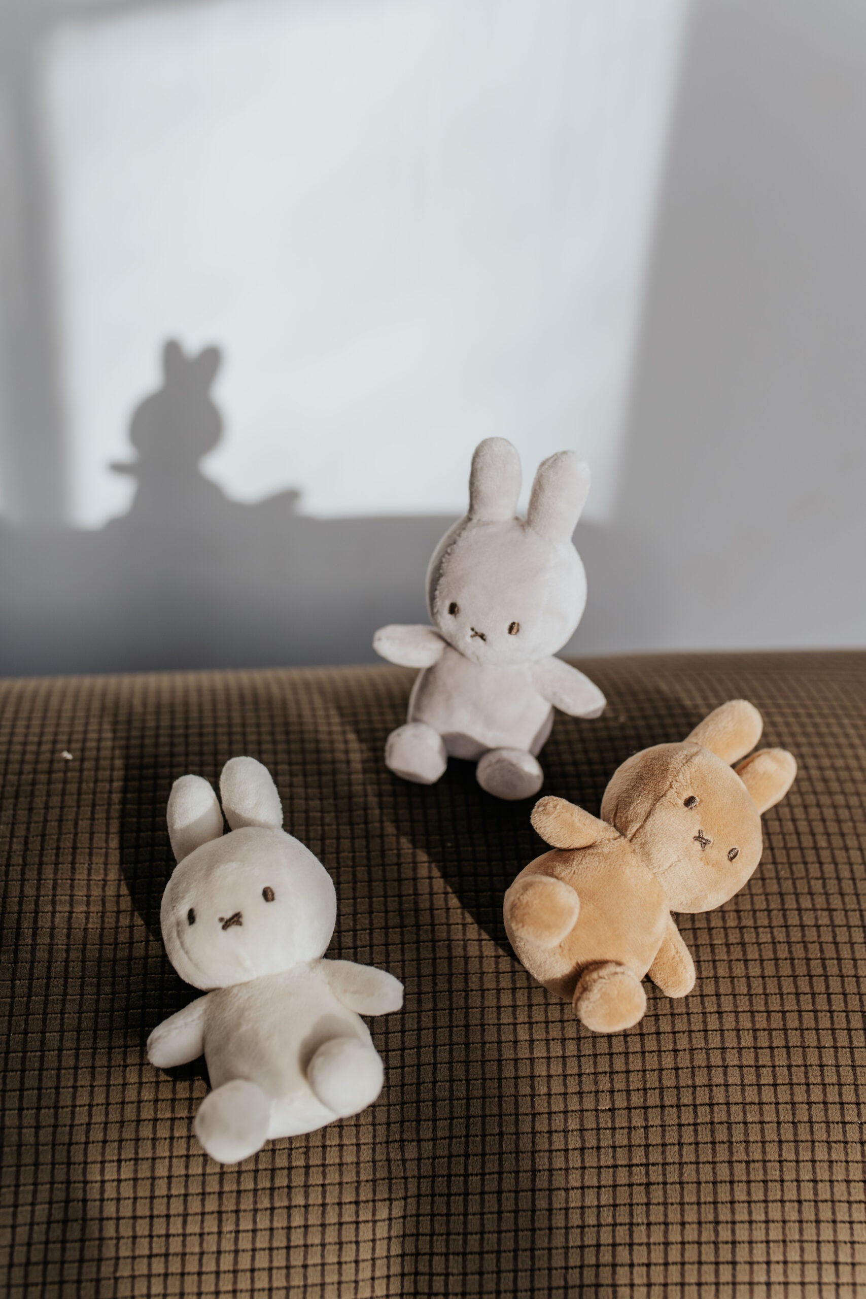 Three Lucky Miffy plush toys from BON TON TOYS X MIFFY rest on a checkered fabric surface. Handcrafted from recycled polyester, the bunnies come in white, light grey, and light brown. The white bunny sits upright while the light grey (Lucky Miffy in Giftbox Grey 10cm) and light brown ones lean to the side. A shadow of one bunny is cast on the wall behind them.