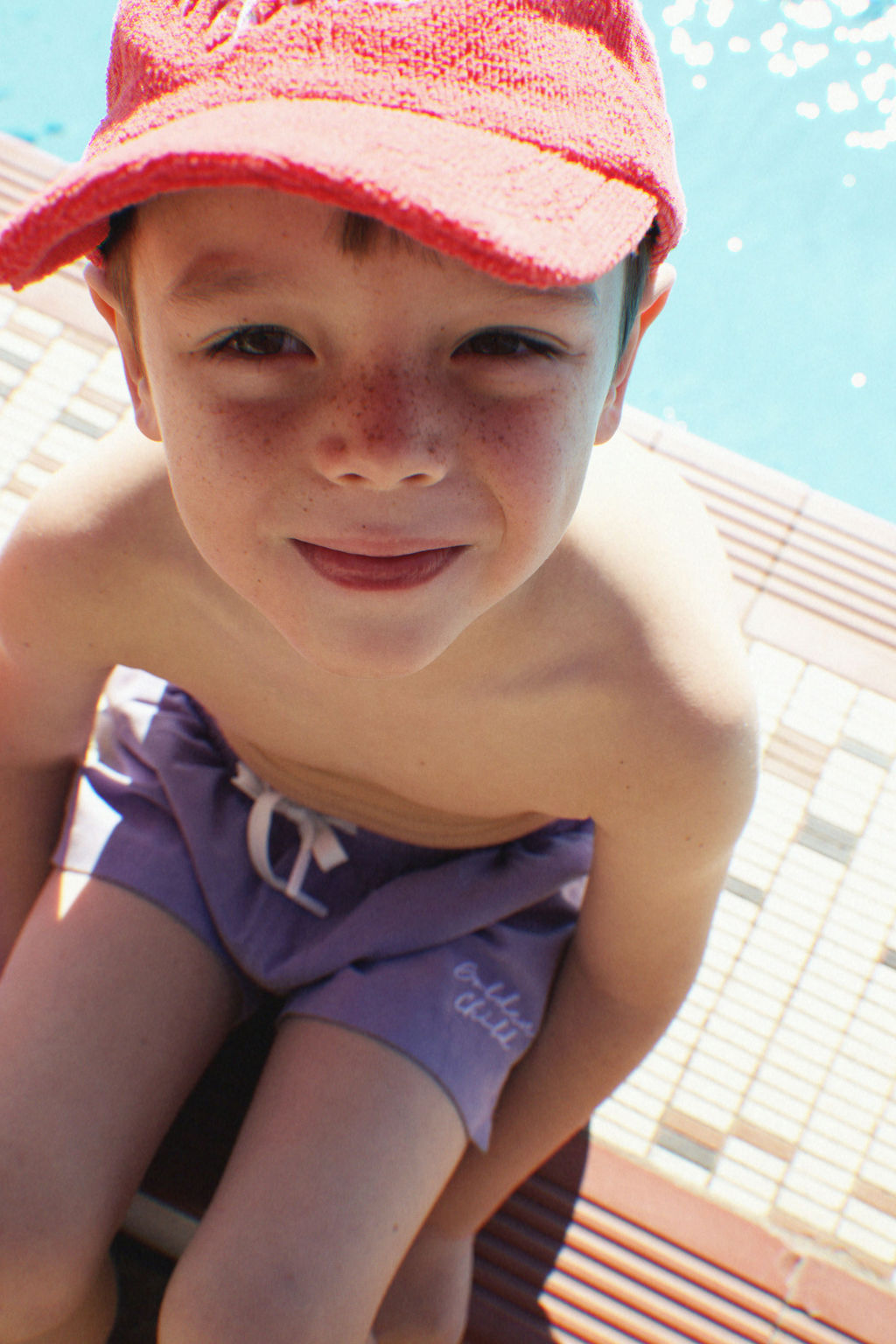 A young boy with freckles smiles at the camera, wearing a Children's Terry Towelling Cap in Racer Red by GOLDEN CHILD along with purple swimming shorts, sitting by the edge of a pool.