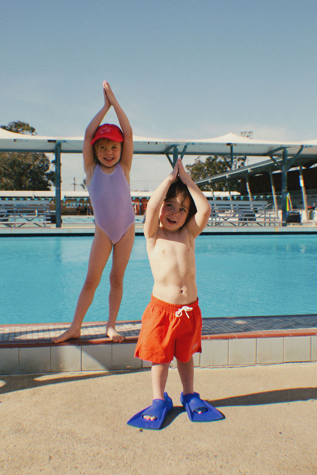 Two spirited children at a pool, one in a purple swimsuit and red cap standing on the edge, the other in GOLDEN CHILD's PRE-ORDER Golden Kids Boardshorts Racer Red featuring an adjustable drawstring waist and quick-dry fabric. Both stand with hands raised playfully against a backdrop of pool and sky.