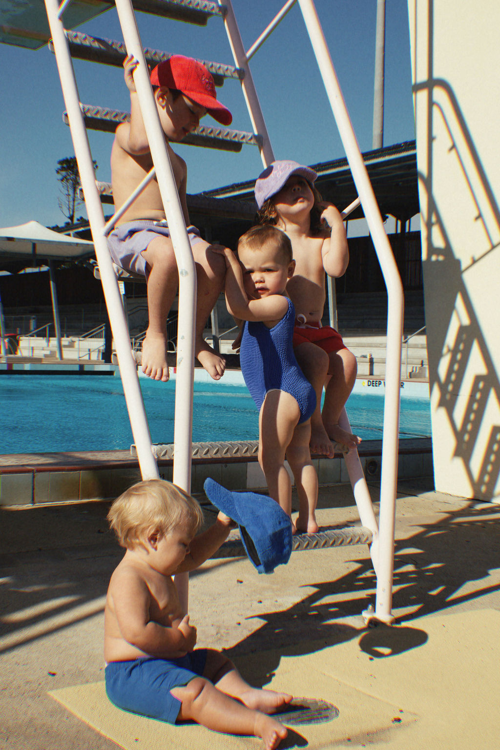 Four children wearing their PRE-ORDER Golden Kids Boardshorts Electric Blue by GOLDEN CHILD are playing on a pool ladder. Two sit on the steps, one hangs from the railing, while another stands on the ground with a blue hat. With quick-dry fabric and an adjustable drawstring waist, they're ready for fun under the sunny sky by the pool.