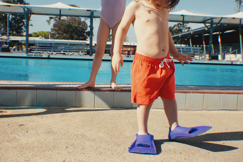 A child in vibrant Golden Child PRE-ORDER Golden Kids Boardshorts Racer Red with an adjustable drawstring waist and blue flippers stands by the edge of a sunny swimming pool. An adult is nearby, partially visible, amidst trees and covered seating glistening under the sun.