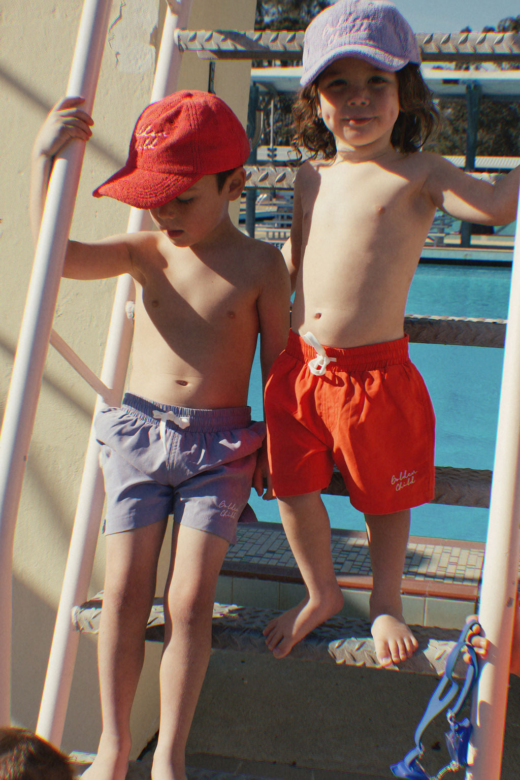 Two children wearing caps and swim trunks stand on a poolside ladder. The boy sports a red terry towelling cap with gray trunks, while the girl flaunts a GOLDEN CHILD Children's Terry Towelling Cap in Grape featuring an adjustable velcro back and red trunks. The shimmering pool water forms their vibrant backdrop.