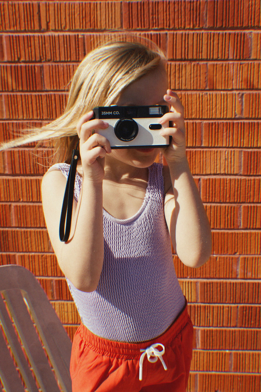 A child with long hair holds a camera up to their face, standing against a red brick wall with a textured surface. They wear the Golden Girls One Piece Crinkle Swimsuit in violet from GOLDEN CHILD, capturing this playful scene and embracing a moment of vibrant nostalgia.