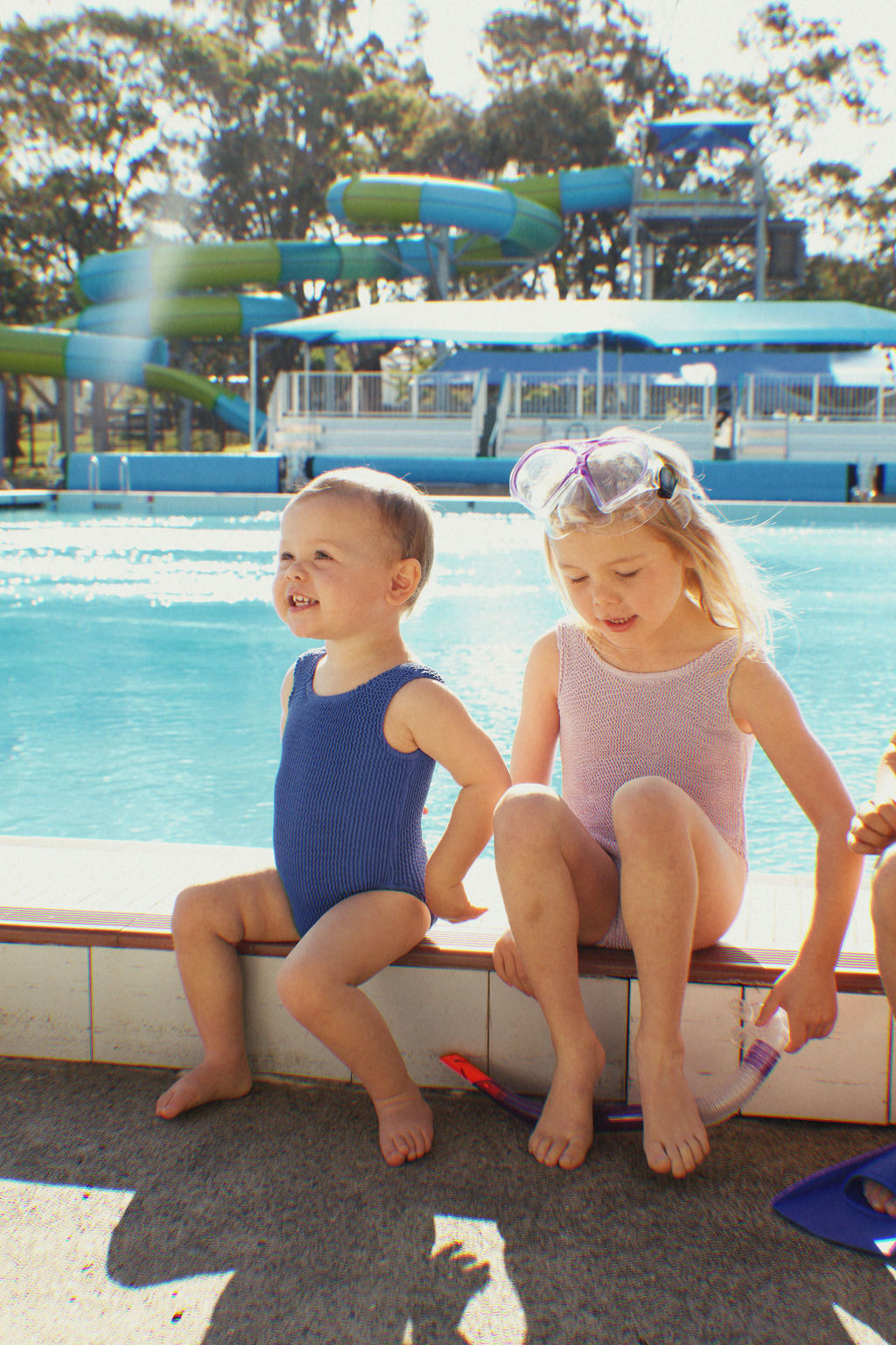 Two children sit by a pool, dressed in swimsuits that exude vibrant nostalgia. The older child is wearing a Golden Girls One Piece Crinkle Swimsuit Violet by GOLDEN CHILD, with goggles perched on her head and a toy in her hand, while the toddler beams with joy. In the background, water slides and trees soak up the sunshine under a clear sky.