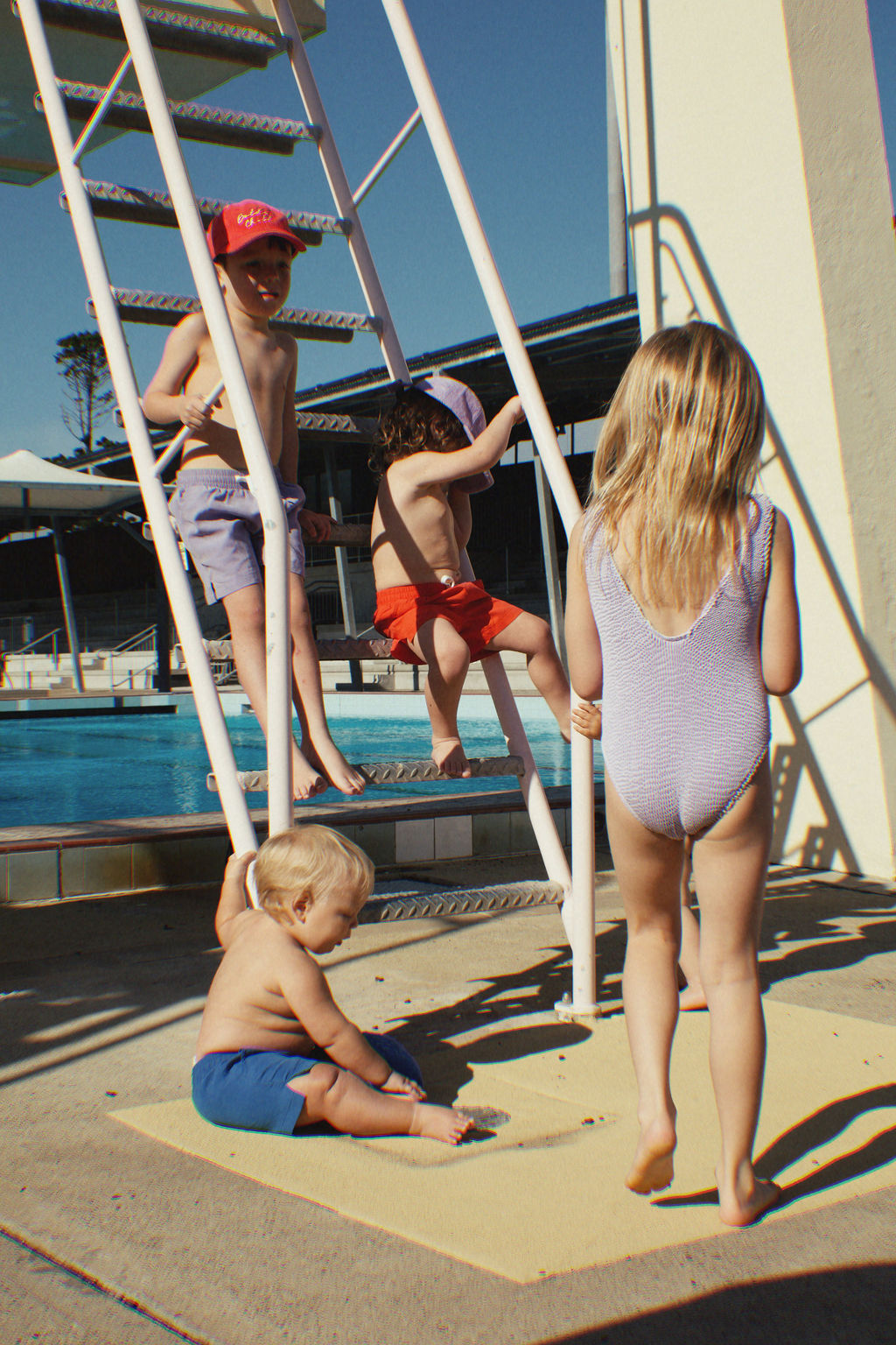 In this sunny scene of children enjoying nostalgic play, a group gathers near a diving board ladder by the swimming pool. One child sits on a mat while others either climb or stand around energetically. Among them, the PRE-ORDER Golden Girls One Piece Crinkle Swimsuit in Violet from GOLDEN CHILD stands out strikingly, with the pool and a small building setting the background.