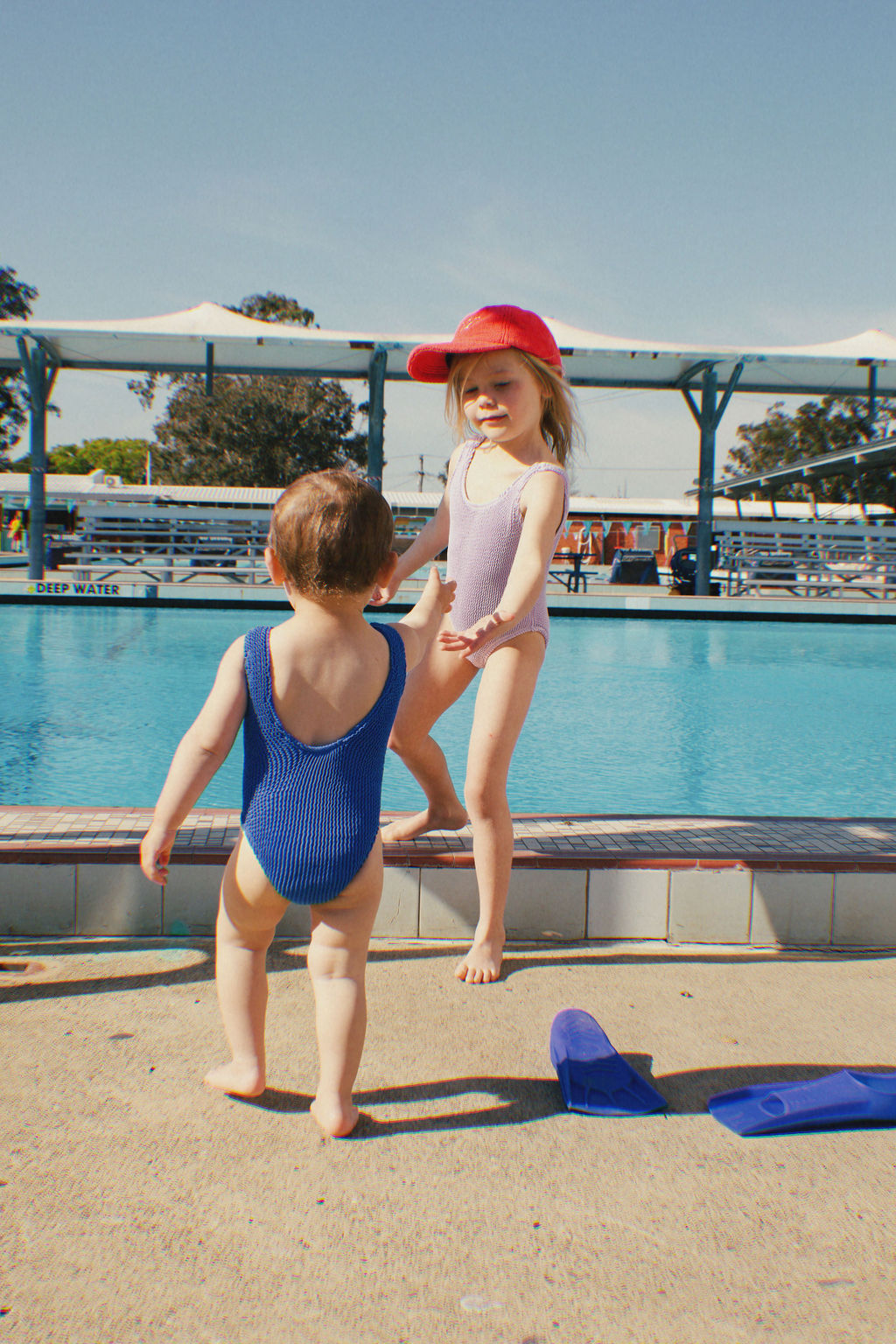 Two young children in swimsuits play by a swimming pool. One child sports a red cap and purple swimsuit made of crinkle swim fabric, while the other is wearing the PRE-ORDER Golden Girls One Piece Crinkle Swimsuit in Electric Blue by GOLDEN CHILD. Nearby are a pair of blue flippers, and the pool area is surrounded by shade structures and trees.