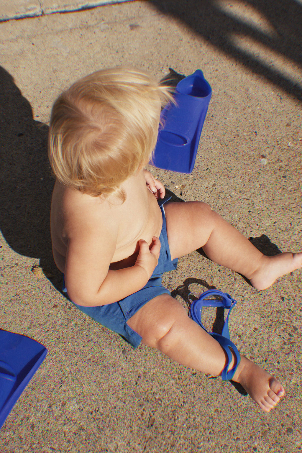 A toddler with blonde hair sits on a concrete surface, sporting Electric Blue Golden Kids Boardshorts by GOLDEN CHILD. The adjustable drawstring waist provides a comfortable fit, and the quick-dry fabric is prepared for water adventures. Nearby, blue swim fins and goggles bask in the sunlight while shadows play across the ground.