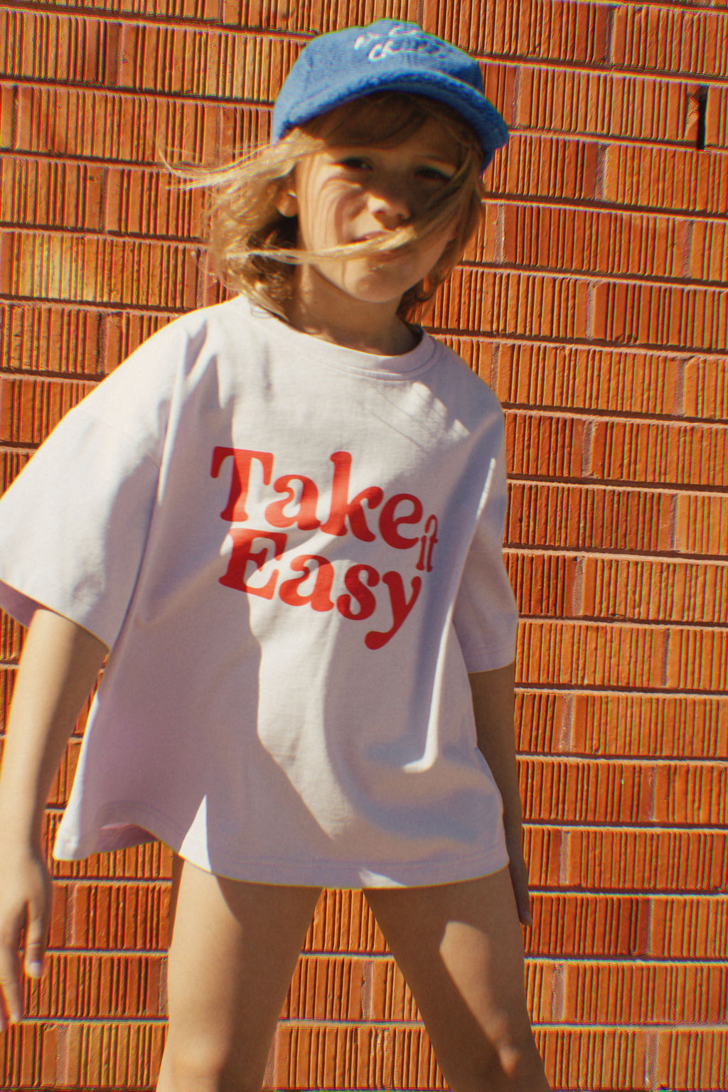 A child wearing the PRE-ORDER The Take It Easy Kids T-shirt by GOLDEN CHILD and a blue cap stands confidently in front of a red brick wall.