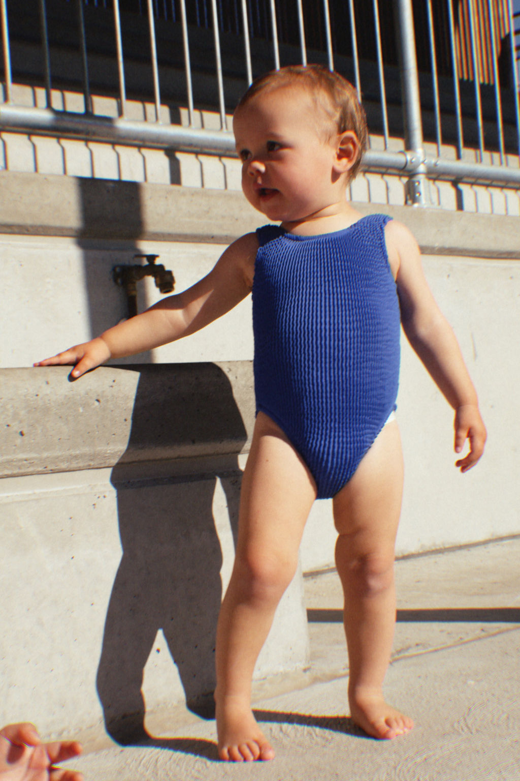 A young child stands barefoot on a sunlit concrete surface wearing the PRE-ORDER Golden Girls One Piece Crinkle Swimsuit in Electric Blue from GOLDEN CHILD. The child leans against a step with one hand, casting a shadow on the ground, while a metal railing and part of a building are visible in the background.