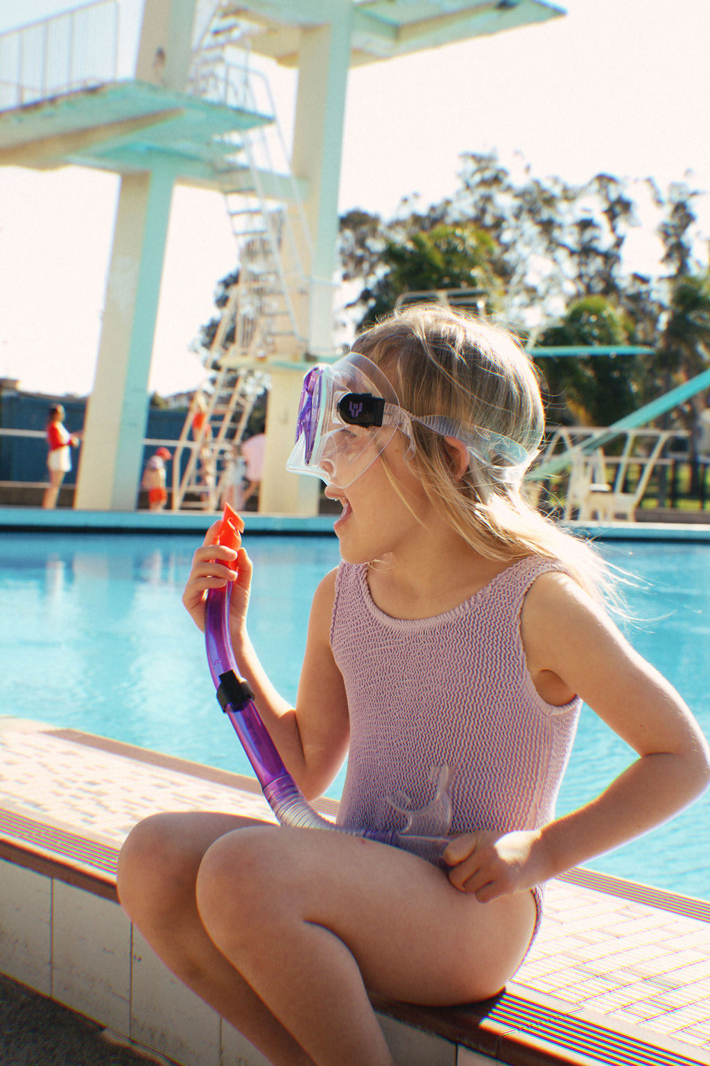 A young girl dressed in a **PRE-ORDER Golden Girls One Piece Crinkle Swimsuit Violet** by **GOLDEN CHILD** sits on the pool's edge, smiling with her snorkel gear. In the background, a diving board and trees bask in vibrant nostalgia under the bright sun.