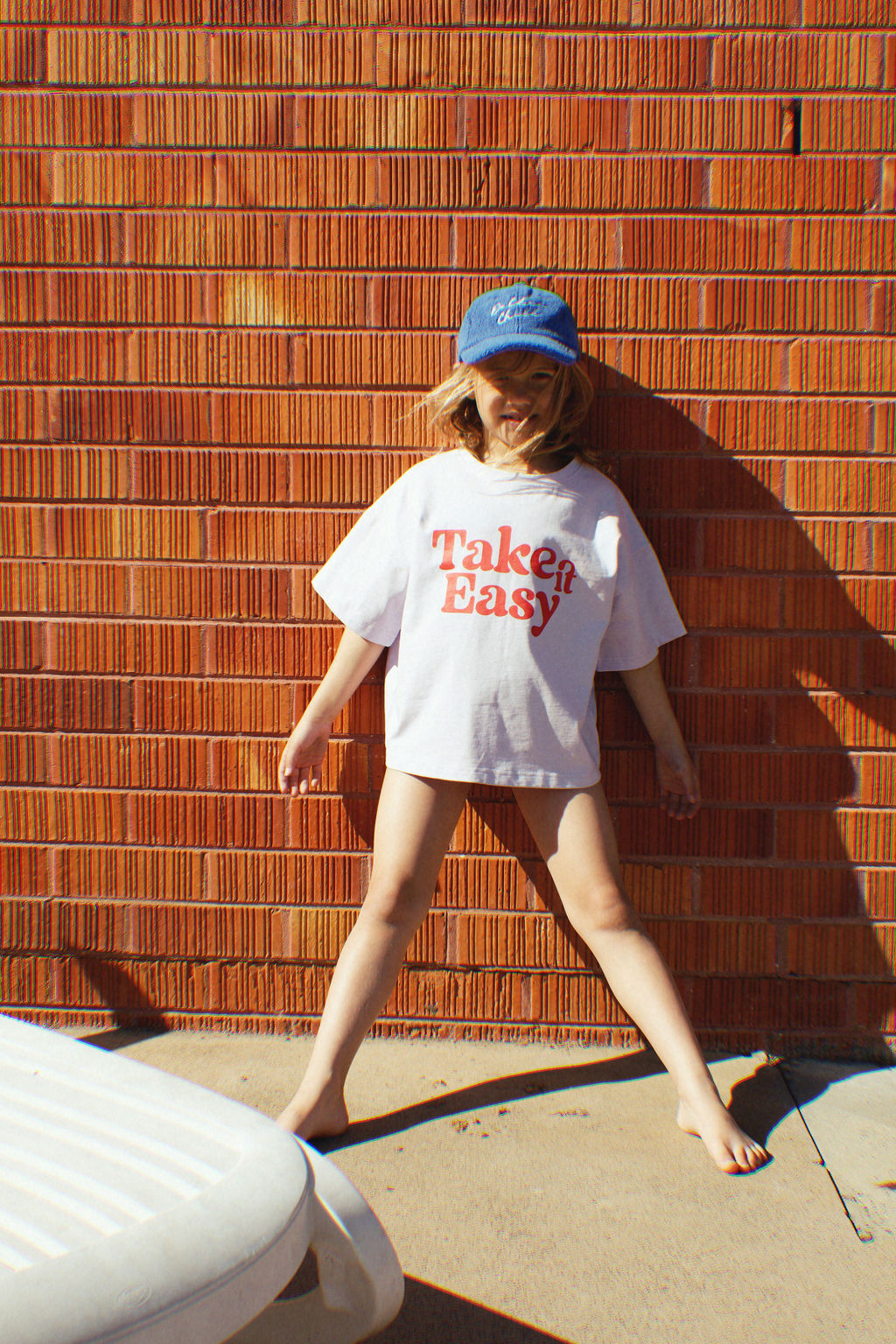 A child playfully stands against a sunlit red brick wall, wearing a retro cool "PRE-ORDER The Take It Easy Kids T-shirt" by GOLDEN CHILD, paired with a blue cap and no shoes. A white plastic chair is partially visible in the foreground.