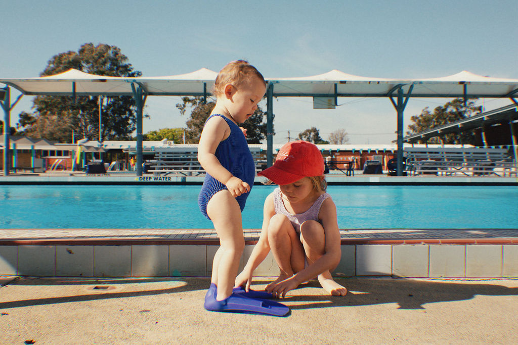 Two young children are by a pool. One child is standing, wearing the PRE-ORDER Golden Girls One Piece Crinkle Swimsuit in Electric Blue from GOLDEN CHILD and matching blue flippers, while the other, in a red cap, is crouched down, helping adjust the flippers. The pool area is sunny and empty.