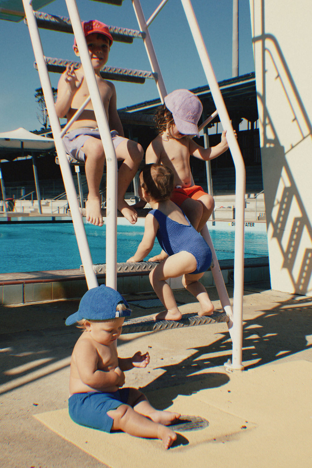 Four children in swimwear enjoy a sunny day at the pool. Two sit on a metal ladder while another climbs. Nearby, a child in a blue hat relaxes on the ground. Their PRE-ORDER Golden Kids Boardshorts in Electric Blue by GOLDEN CHILD, featuring an adjustable drawstring waist, provide comfort and quick-drying fun all day long.