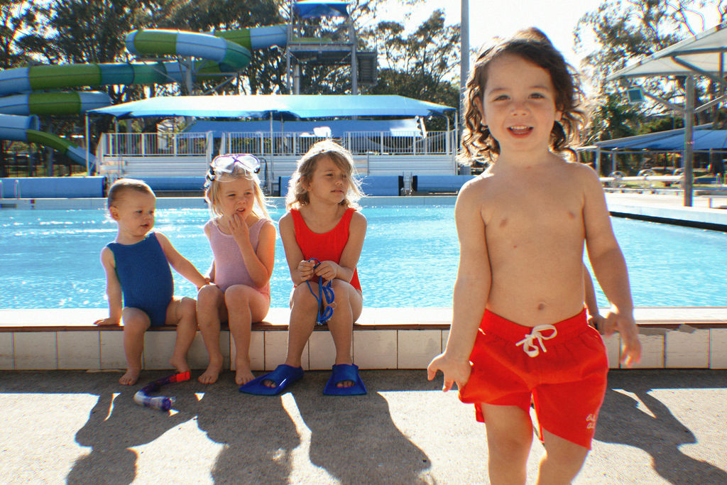 Four children are by a pool; three are sitting at the edge in swimsuits, and one child is walking towards the camera wearing Golden Kids Boardshorts Racer Red with an adjustable drawstring waist. In the background, a waterslide and trees create the impression of an outdoor water park, perfect for enjoying quick-dry fabric fun.