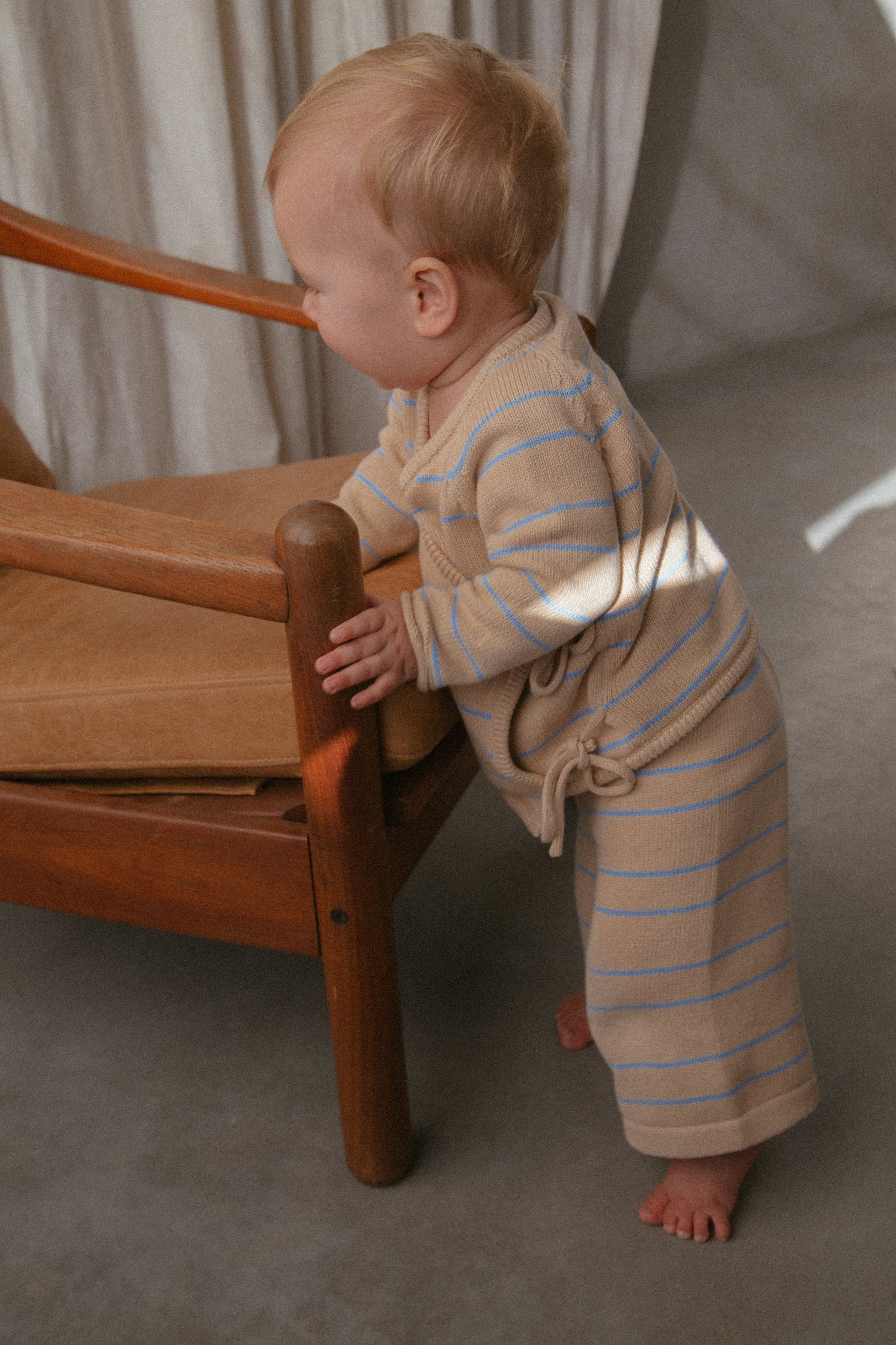 A small child with light blonde hair, dressed in MILKY DESIGNS' Essential Wrap Cardigan Blue Stripe made of 100% cotton, stands while holding onto a wooden armchair with a tan cushion. The child's gaze is directed downward, and the background includes soft, neutral-colored curtains.