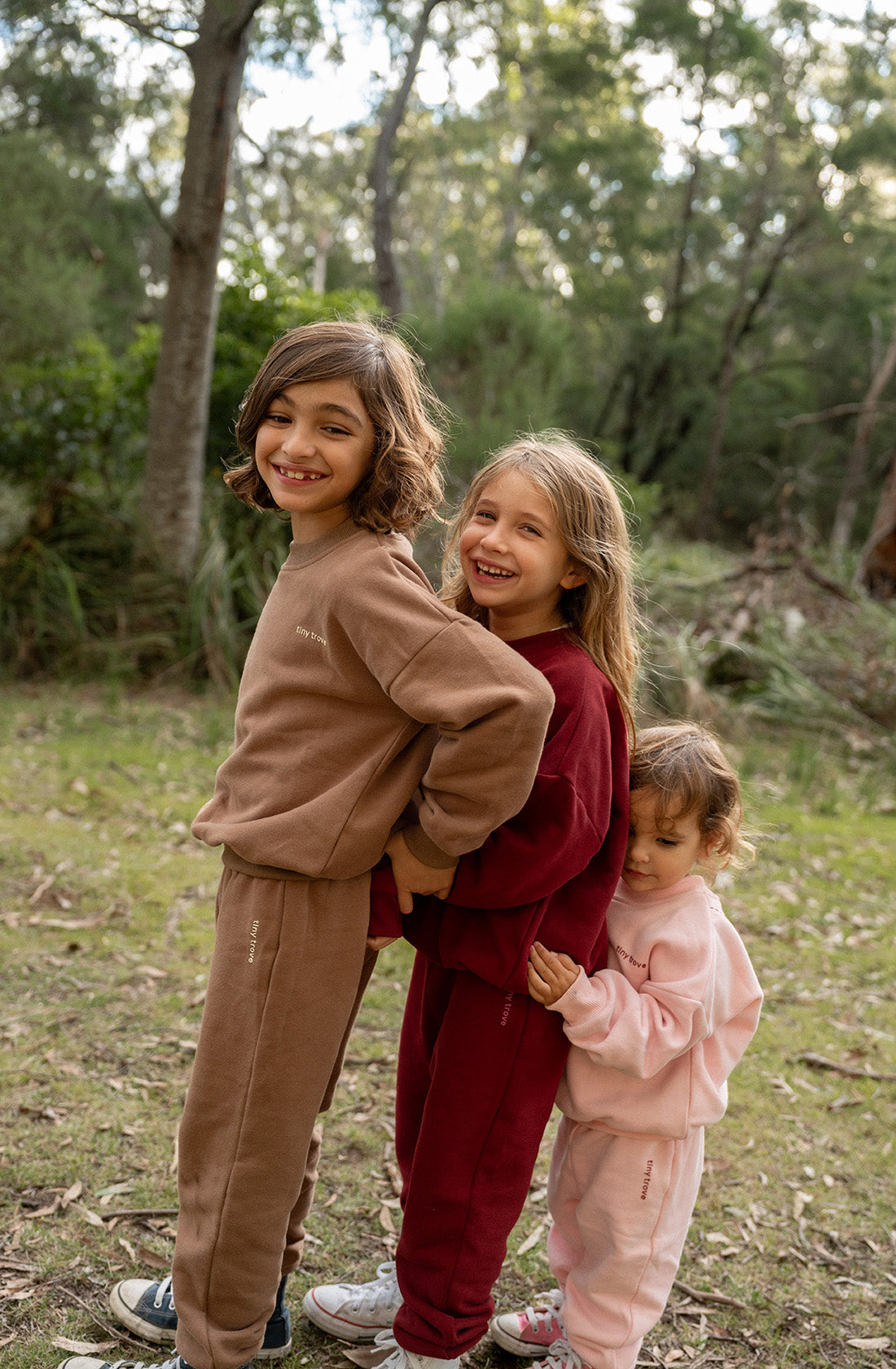 Three children standing outdoors in a line, smiling and dressed in different colored sweatsuits—including the TINY TROVE Woodie 3D Logo Tracksuit Mocha. Made from soft cotton jersey with subtle 3D logos, they are surrounded by a forested area with trees and greenery, enjoying a joyful moment together.