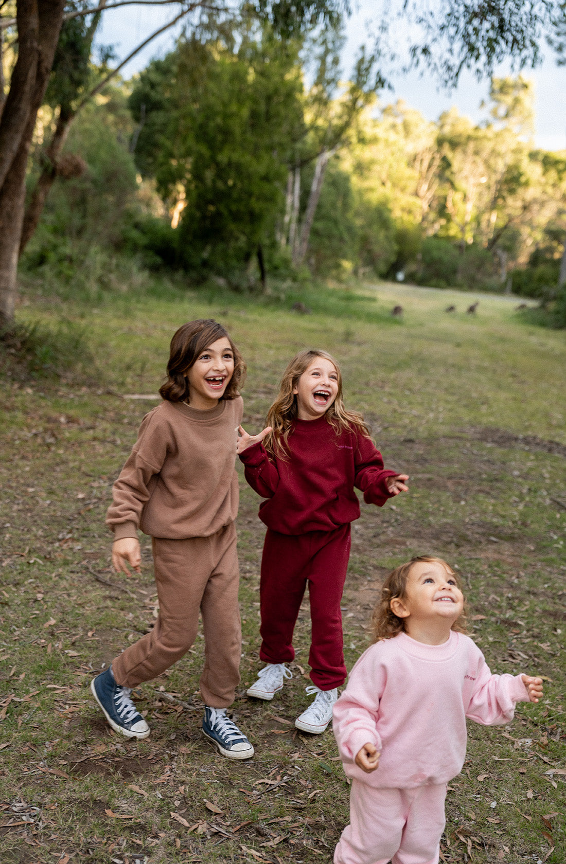 Three young children are playing outdoors in a wooded area. They are all smiling and looking up, appearing joyful. The oldest child wears a TINY TROVE Woodie 3D Logo Tracksuit in mocha, the middle child wears a maroon outfit, and the youngest child wears a light pink outfit. Trees and grass surround them.