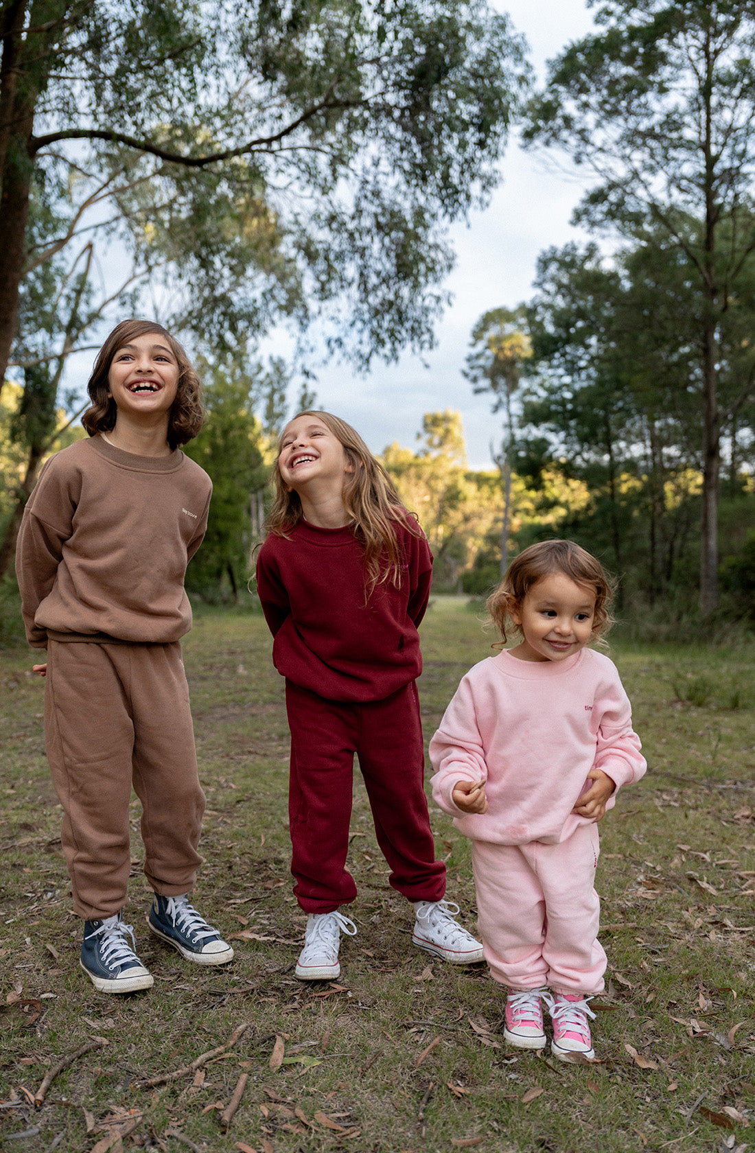 Three children are standing outdoors in a grassy, wooded area. They are all wearing soft cotton jersey tracksuits; the child on the left is dressed in a TINY TROVE Woodie 3D Logo Tracksuit Mocha, the child in the middle is wearing a maroon tracksuit, and the child on the right is sporting a pink tracksuit. They are smiling and looking happy.