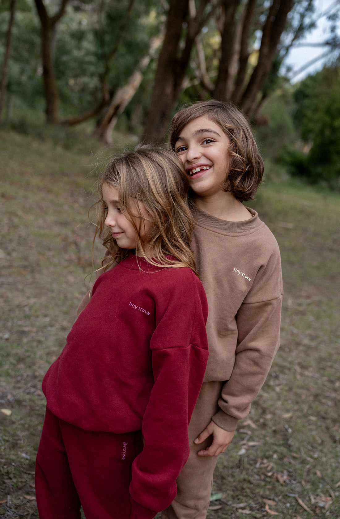 Two children stand in a grassy area with trees. The child on the left, dressed in a maroon fleece cotton outfit, looks down, while the child on the right wears the Woodie 3D Logo Tracksuit Mocha by TINY TROVE, smiles, and leans slightly toward the other. Both outfits feature "TINY TROVE" printed on them.