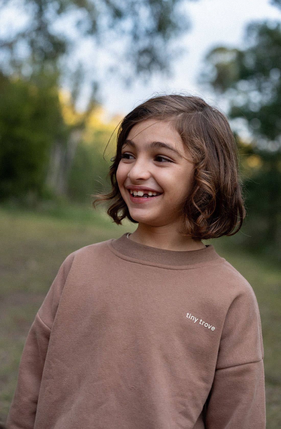 A young child with shoulder-length brown hair is standing outdoors, smiling broadly. The child is wearing a mocha-colored sweatshirt from the TINY TROVE Woodie 3D Logo Tracksuit collection, crafted from soft cotton jersey. There are trees and foliage in the blurred background.