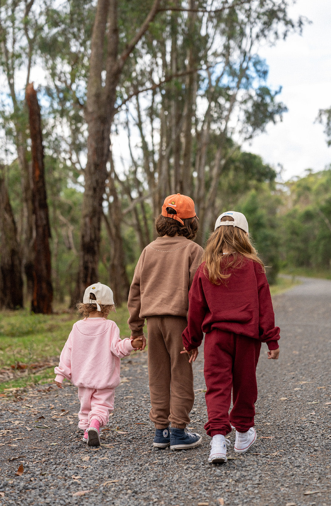 Three children, each wearing matching baseball caps and colorful tracksuits made of soft cotton jersey from TINY TROVE, walk hand-in-hand down a gravel path surrounded by tall trees. The smallest child wears a pink outfit, while the older two wear the Woodie 3D Logo Tracksuit Mocha in brown and maroon outfits, respectively.