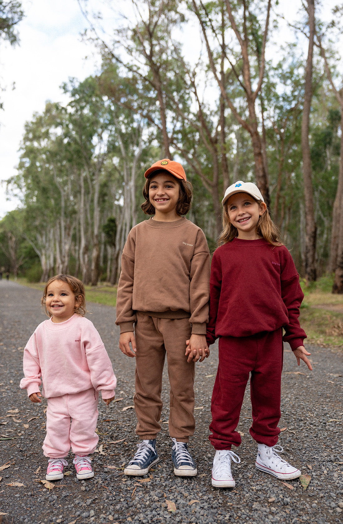 Three children stand outdoors on a gravel path surrounded by trees. The smallest child on the left wears a pink soft cotton jersey outfit, the middle child wears a mocha-colored Woodie 3D Logo Tracksuit by TINY TROVE paired with an orange cap, and the tallest child on the right wears a maroon outfit with a 3D logo and white cap. All are smiling.
