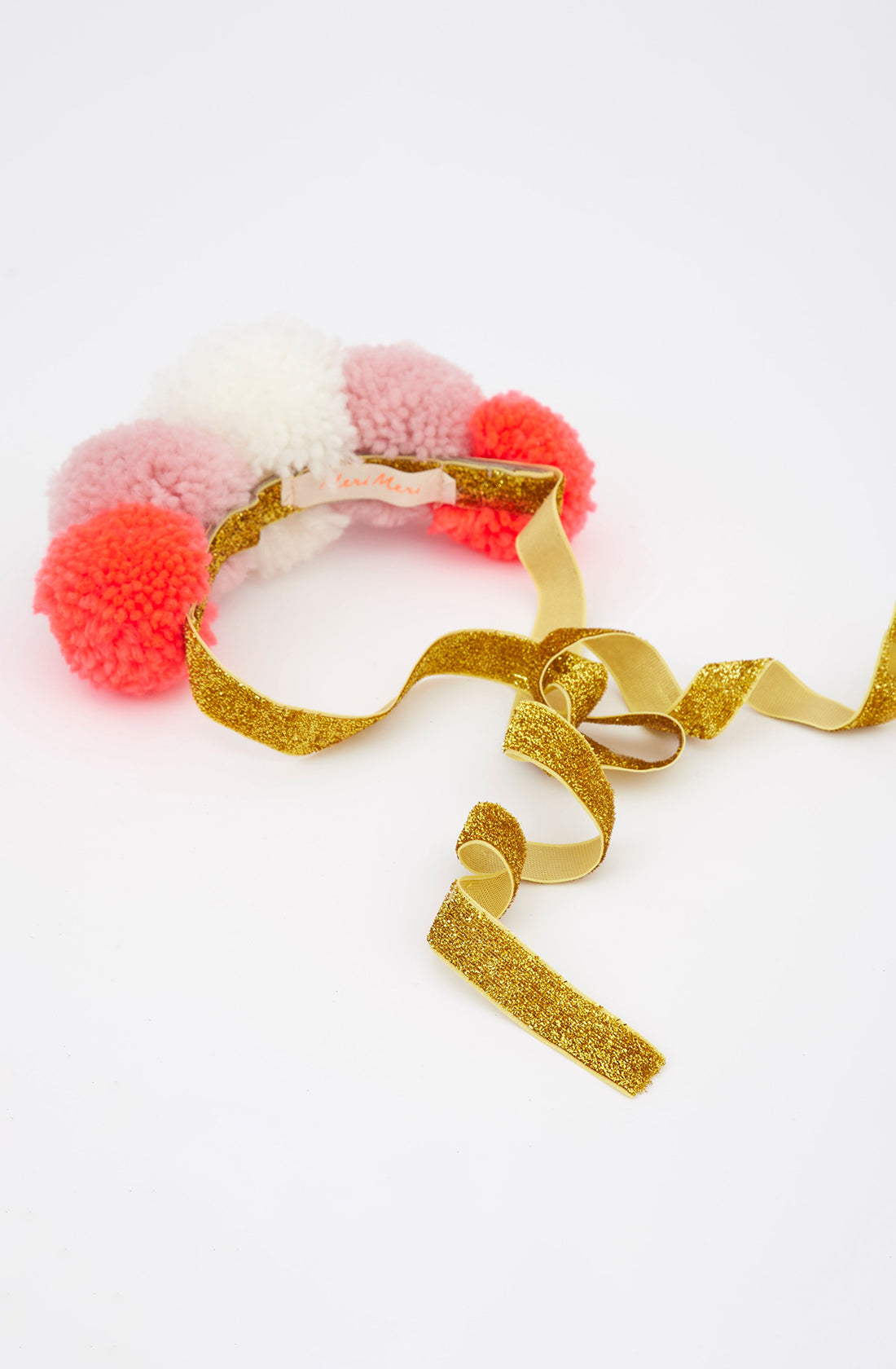 A decorative MERI MERI Pom Pom Crown, featuring pink, white, and red pom-poms, lies on a white surface. Attached to the crown is a long glittery gold ribbon that cascades loosely toward the front—perfect for any dressing-up costume.