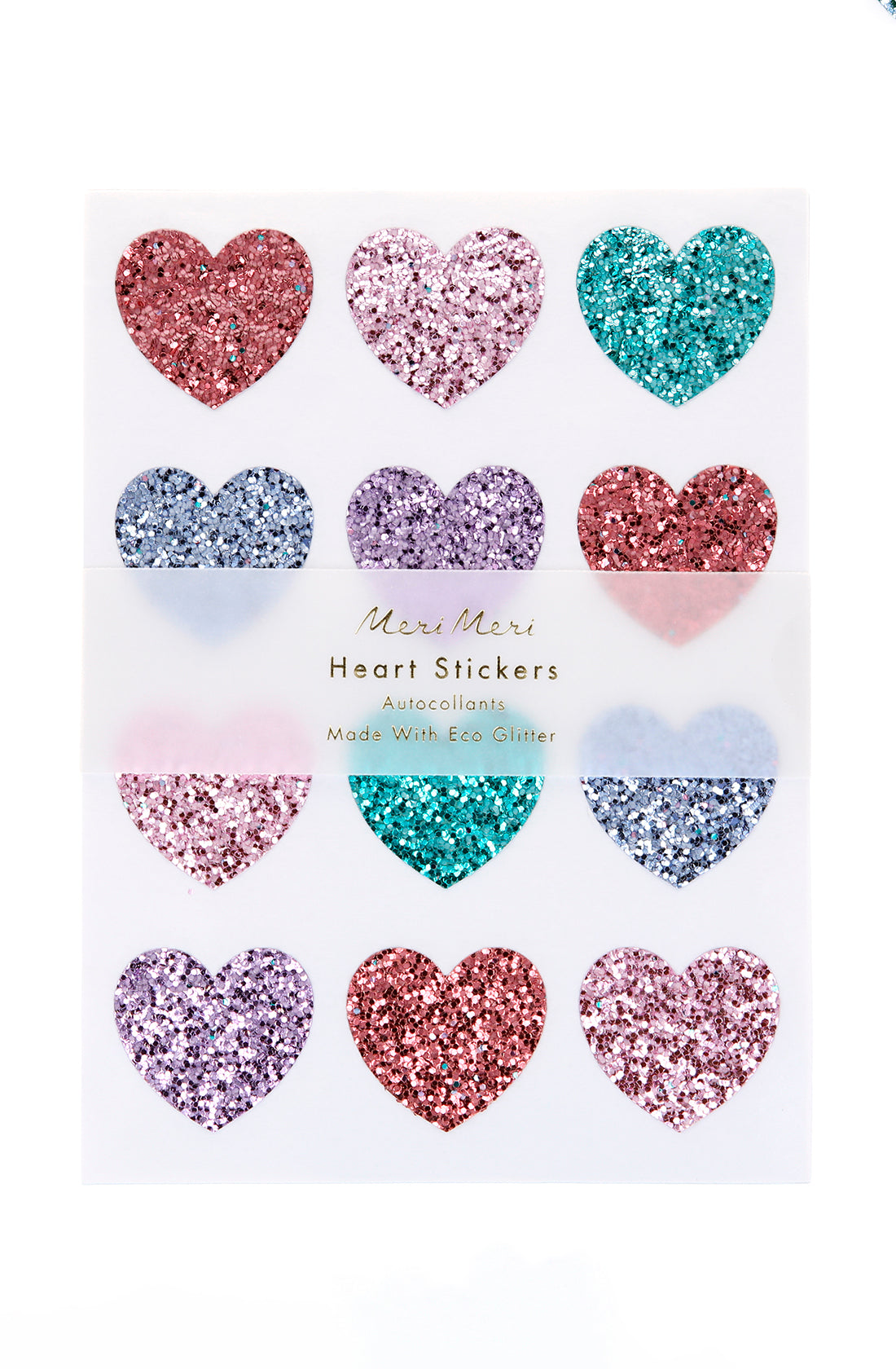 A sheet of twelve heart-shaped stickers arranged in a 3x4 grid. Each heart, coated with eco glitter, showcases vibrant colors like pink, red, purple, blue, and green. The label reads "Rainbow Glitter Heart Stickers by MERI MERI" and notes they are perfect for Valentine's Day.