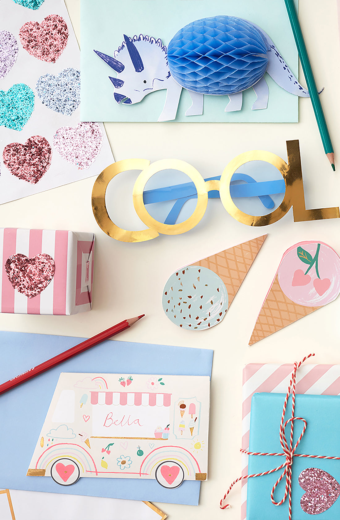 A flat lay of various colorful stationery items including Rainbow Glitter Heart Stickers by MERI MERI, a light blue honeycomb triceratops, gold-rimmed glasses with "COOL" written on them, ice cream cone labels, a pink-striped box, pencils, and a pastel ice cream truck card labeled "Bella".