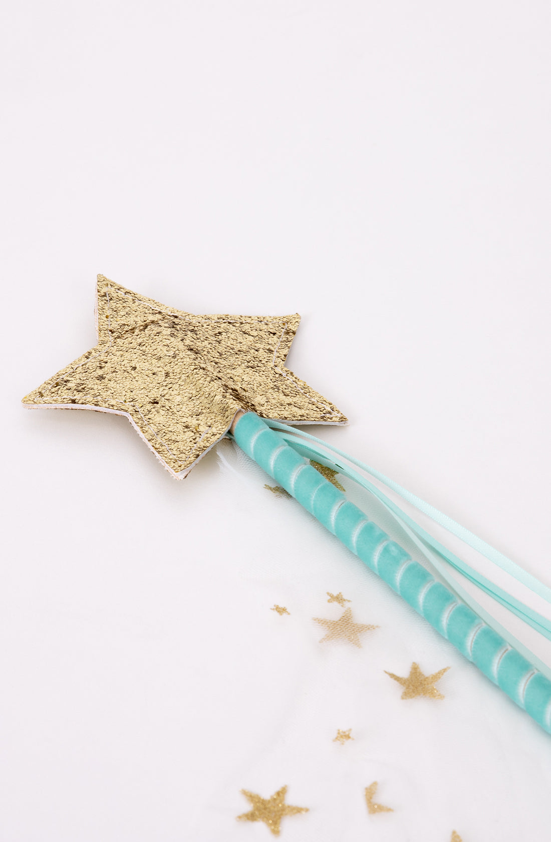 The Tulle Star Wand Mint by MERI MERI features a glittery mint star on top, attached to a blue handle adorned with light blue ribbon streamers, perfect for any fairy or princess. The wand is resting on a white surface, with small gold star-shaped confetti scattered around it, making it an enchanting addition to any dress-up box.