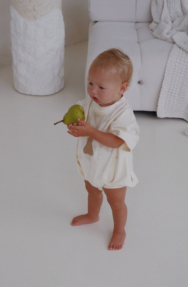 Baby standing holding a pear while wearing the Pea rromper