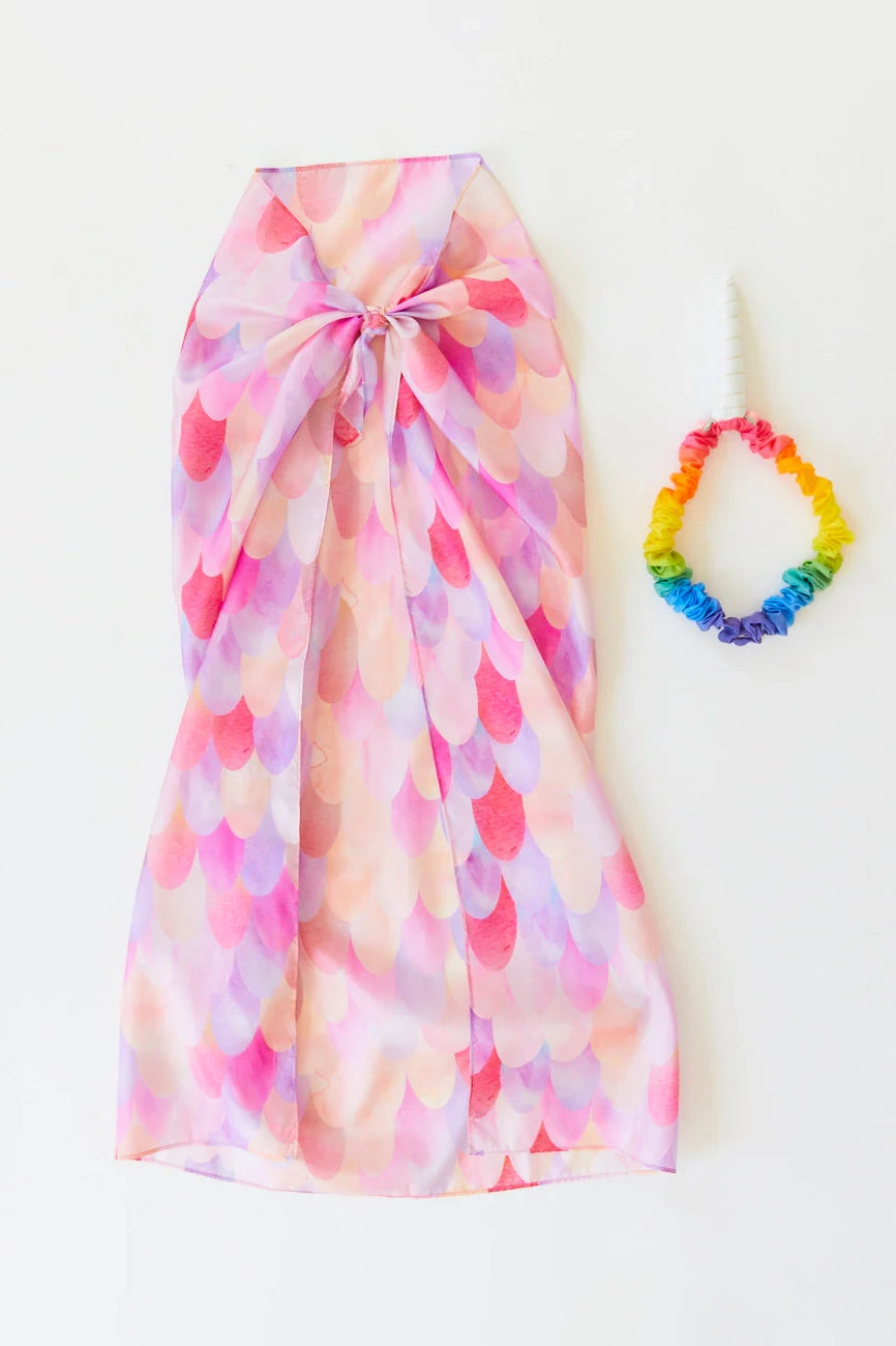 A Pink Unicorn Playsilk-inspired dress featuring a multicolored, scalloped pattern with a halter neck and a large bow on the back, complemented by a rainbow scrunchie, both elegantly displayed on a white background.