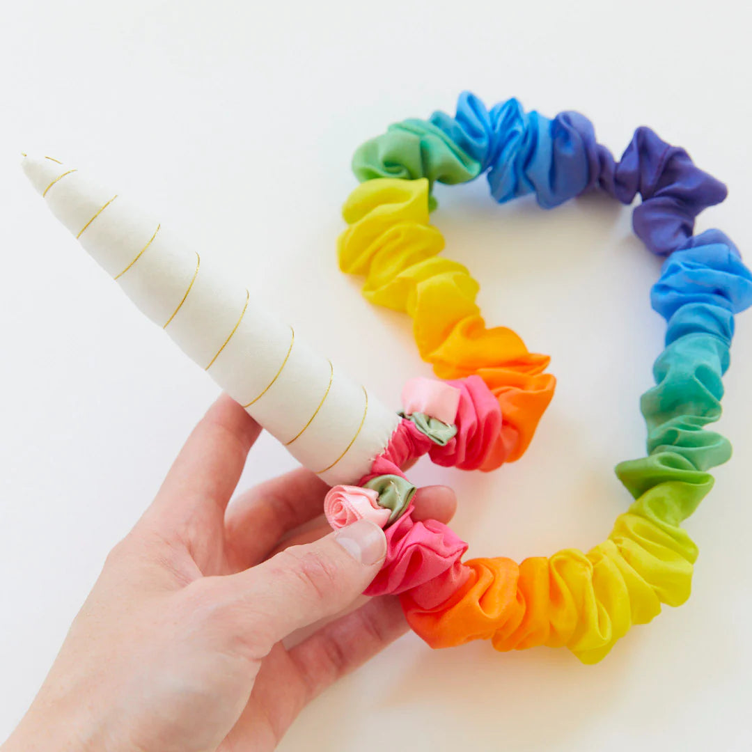 A hand holds a vibrant SCRUNCHIE-style Rainbow Unicorn Headband by SARAH'S SILKS. The white horn, adorned with gold spiral details, is ideal for dramatic or dress-up play and is attached to a band that radiates colors from green to purple.